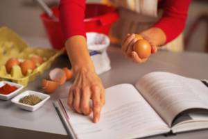 Donna che cucina leggendo il libro delle ricette tenendo in mano un uovo