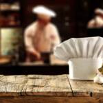 In primo piano un cappello da chef, sullo sfondo lo chef in cucina