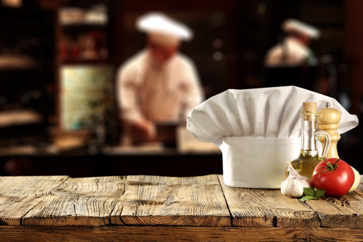 In primo piano un cappello da chef, sullo sfondo lo chef in cucina