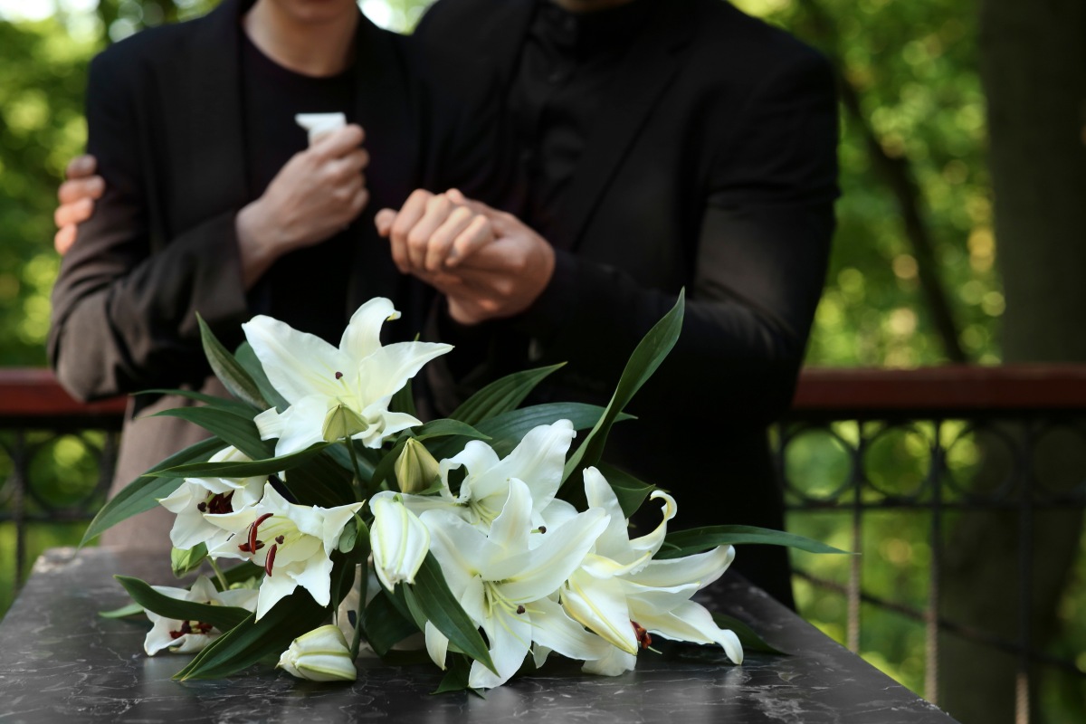 Due persone accostate a una tomba con gigli bianchi in primo piano.